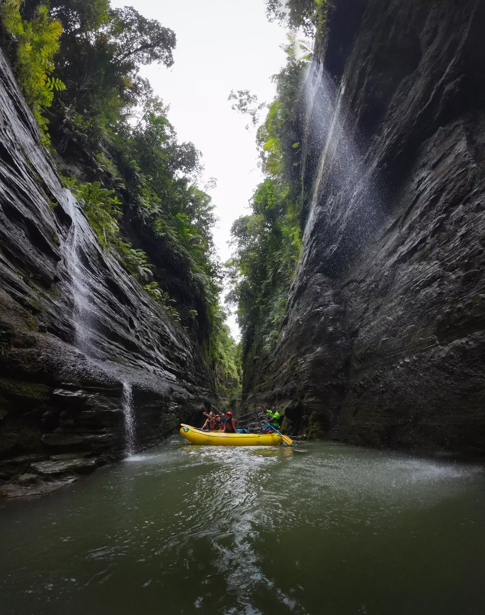 A yellow inflated raft filled with people wearing helmets, life jackets and carrying paddles, travels down a waterway along side two massive rock formations.