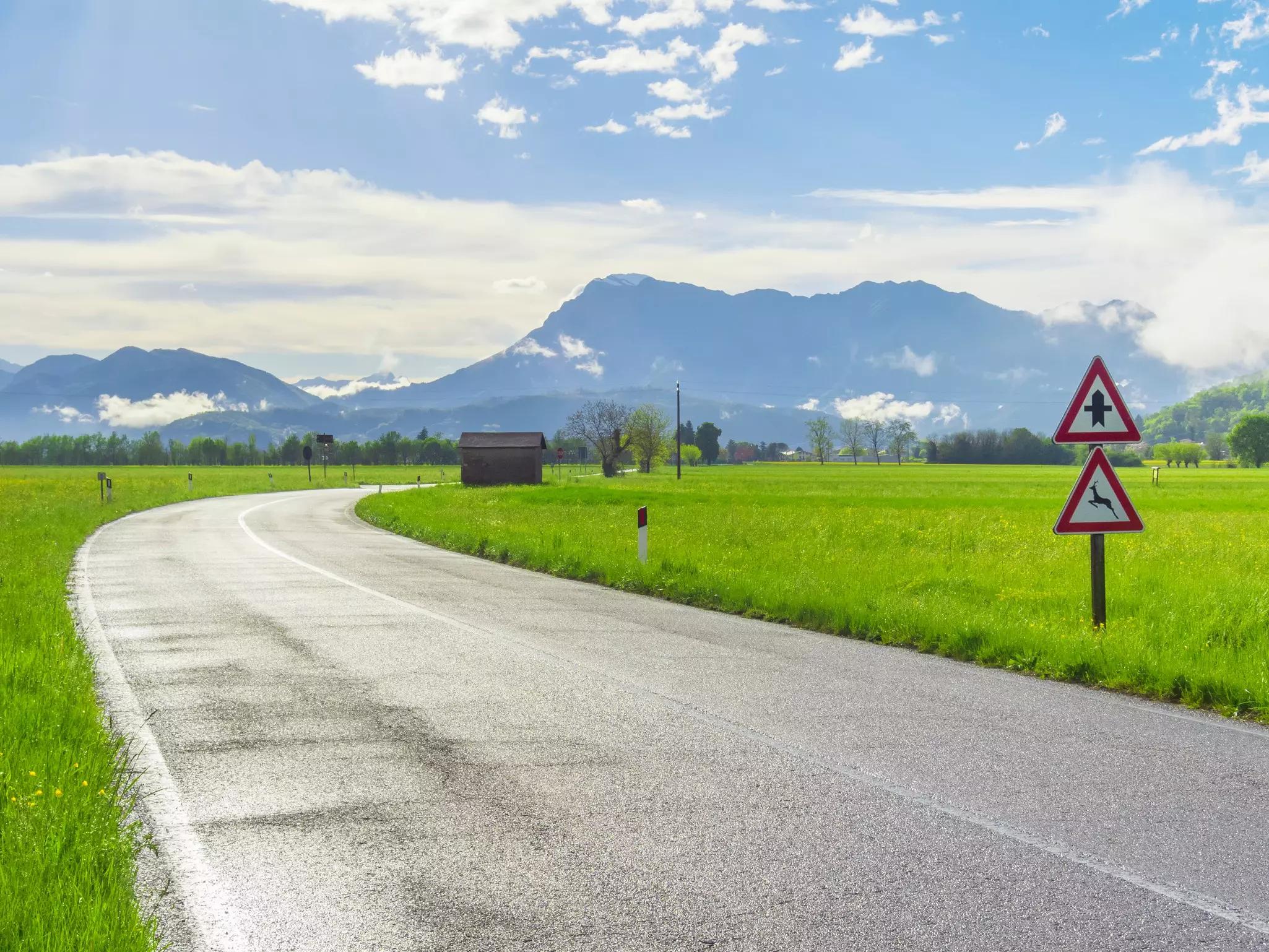 Wet asphalt road after rain in the countryside with road signs, green grass around and mountains on the horizon.