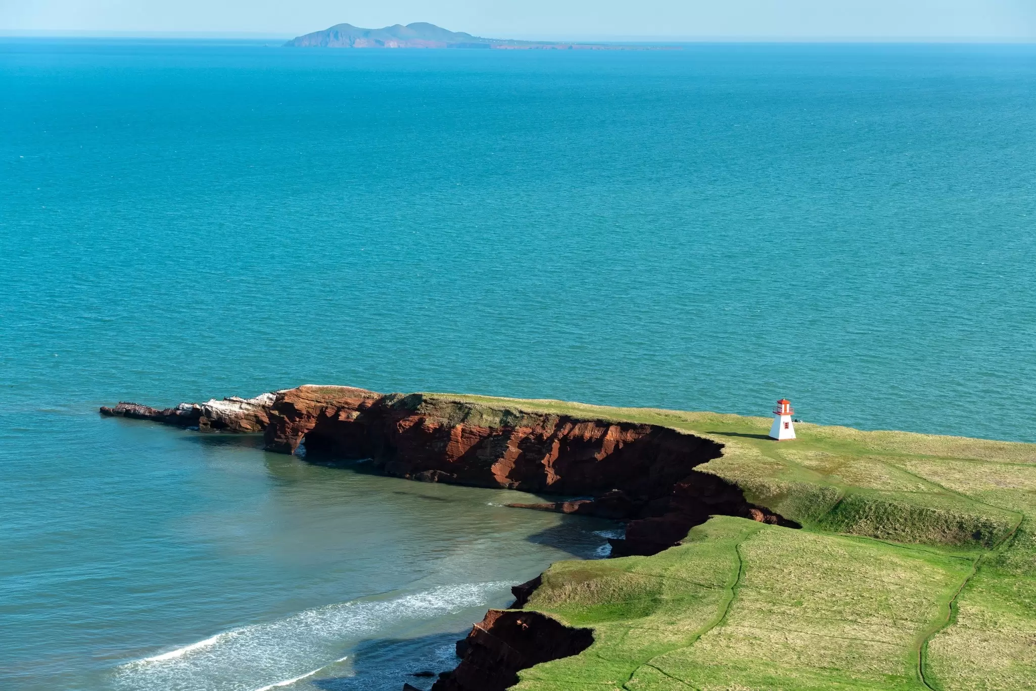 An aerial view of a white lighthouse at the base of a cape with steep red cliffs on an island in the ocean, Îles-de-la-Madeleine, Québec, Canada.