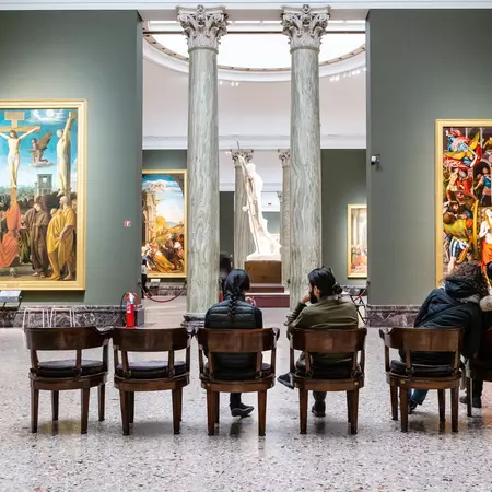 Two people sit in wooden chairs in an ornate Italian art gallery. 