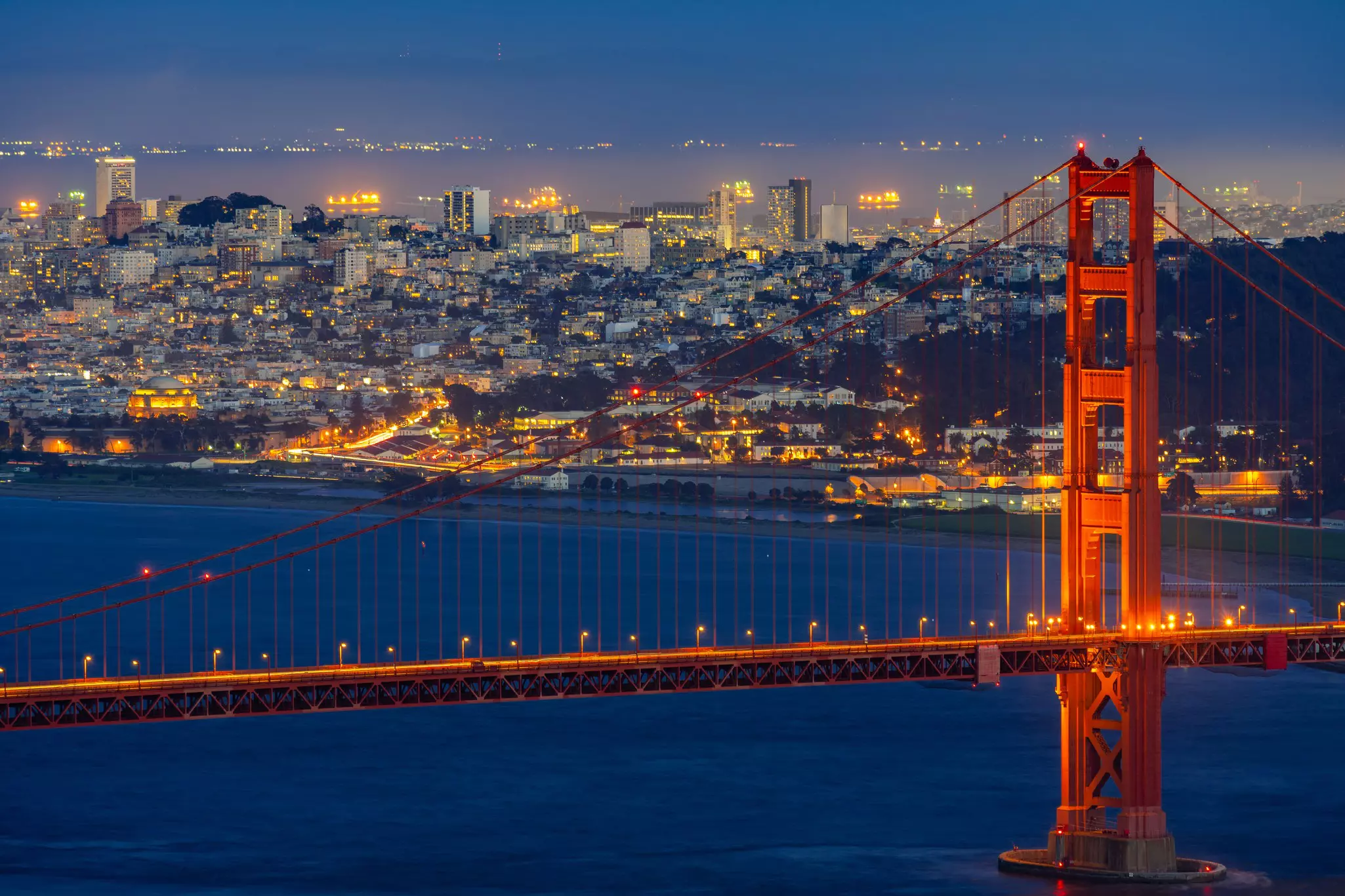 The illuminated Golden Gate Bridge at night over deep blue water with city buildings and homes in the background