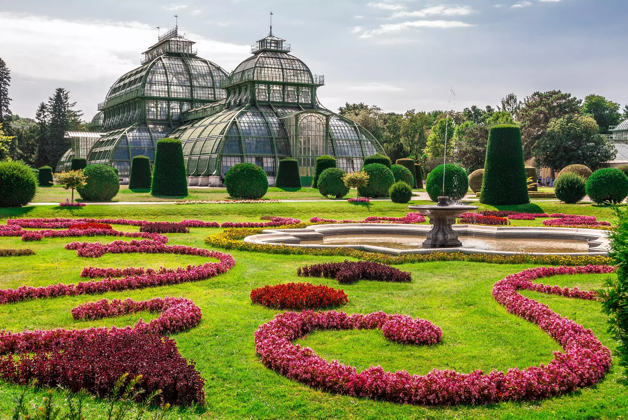 The lush grounds of Palmenhaus, a large greenhouse in the garden of Schloss Schönbrunn Palace, with bright pink, red and burgundy flowers in swirling layouts.