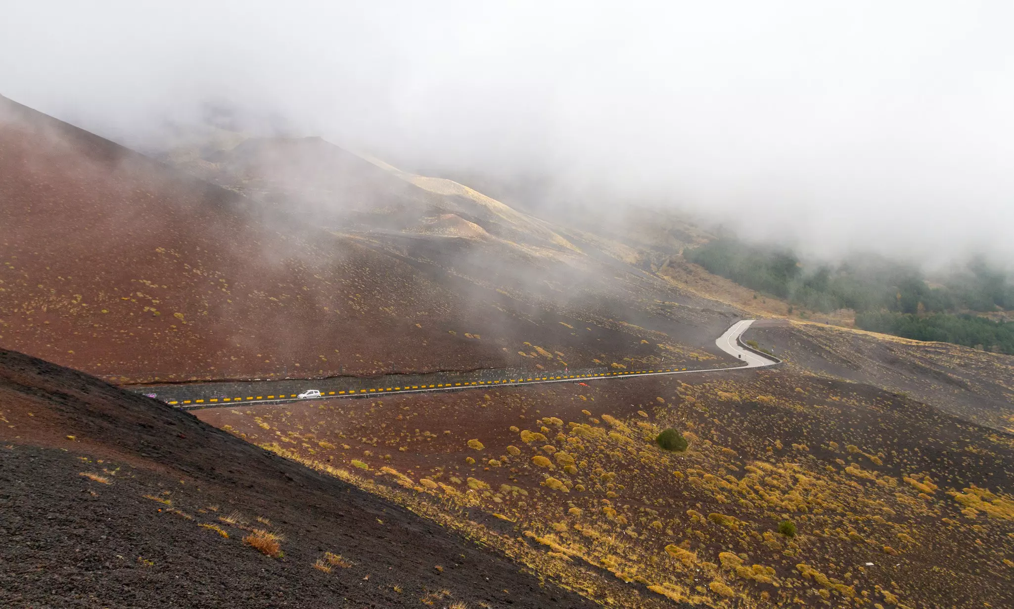 A car drives on a windy road along the ridge of a volcano. Mist shrouds the scene.