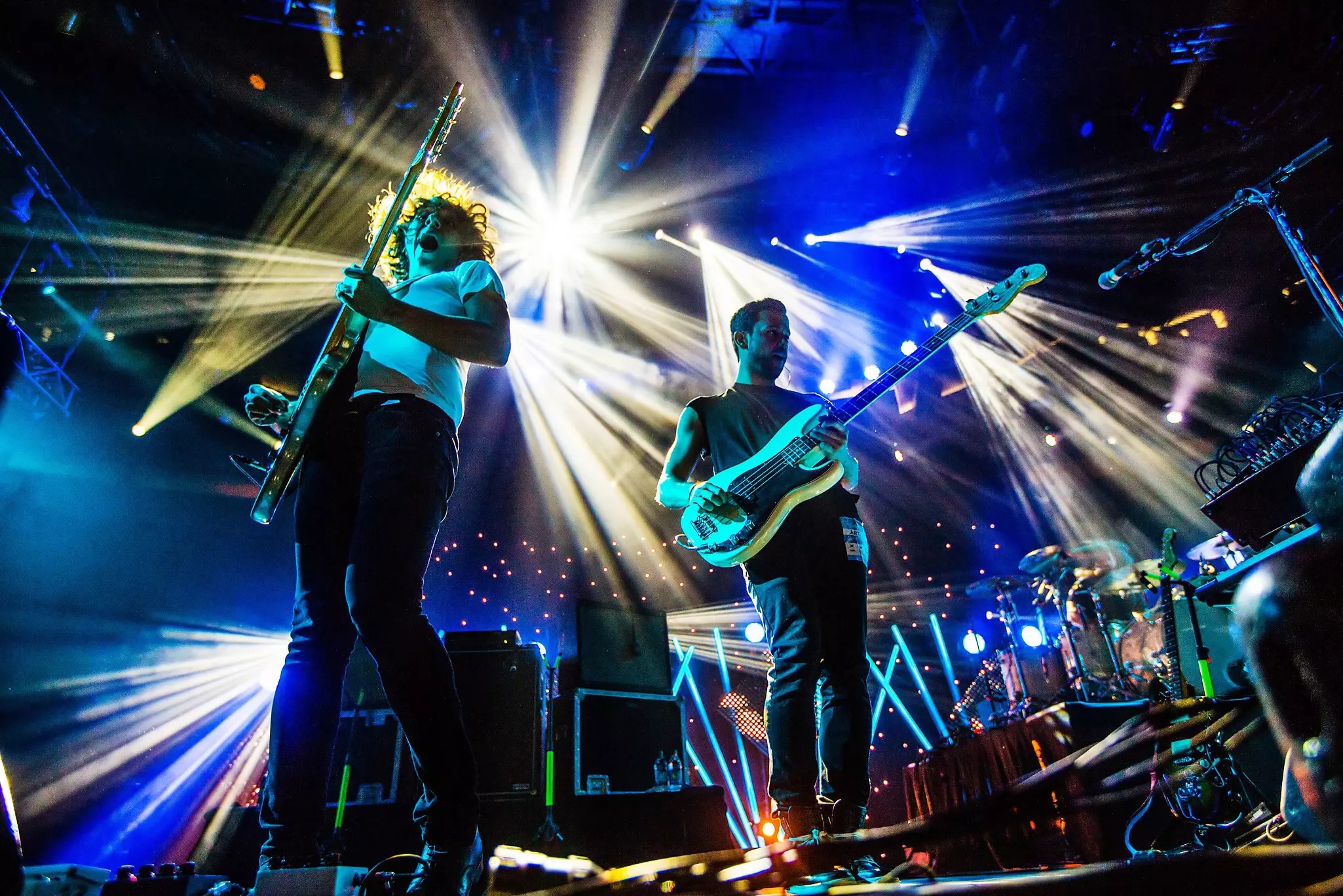 Two guitarists perform live on stage surrounded by spotlights in white and blue.