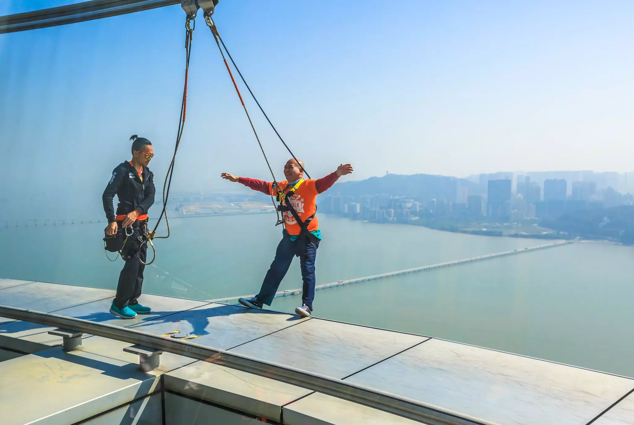 Two people on the ledge during the popular Skywalk at Macau Tower. One of them is leaning back with open arms and laughing.