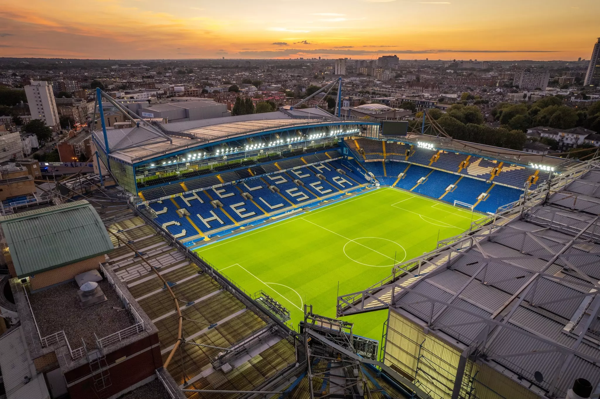 A central grass football pitch in a floodlit stadium surrounded by blue fold-down seats