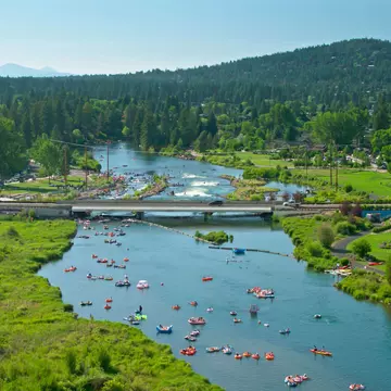 Two people are seen paddleboarding on a deep blue river from a vantage point far above on a rocky outcropping near Bend, Oregon