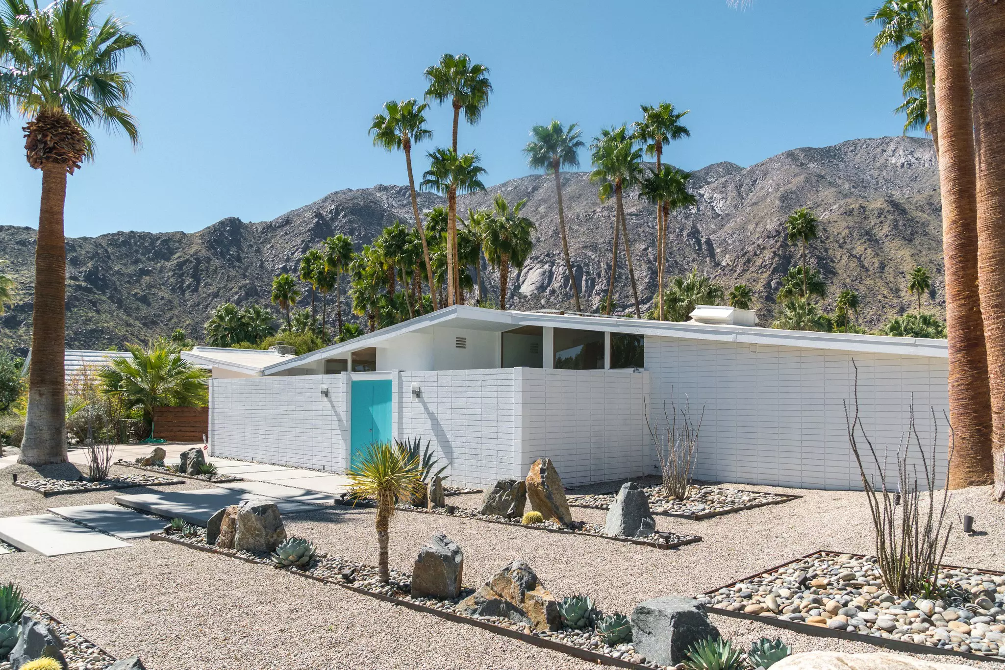 A white, mid-century-modern house sits on a street in a desert city, framed my palm trees. Rocky hills are visible in the distance.