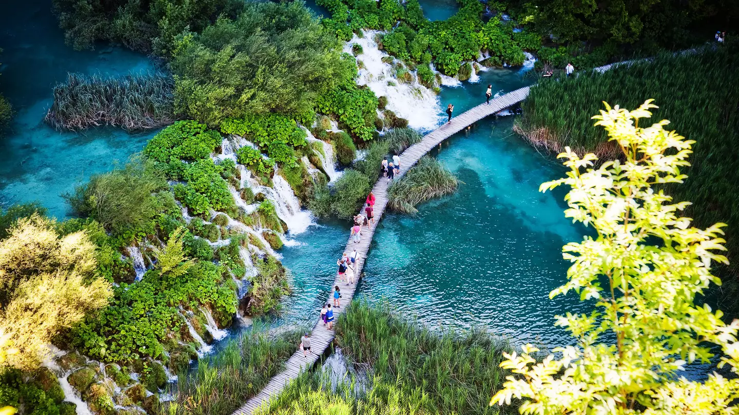 People walk on a pathway over water past a series of waterfalls.