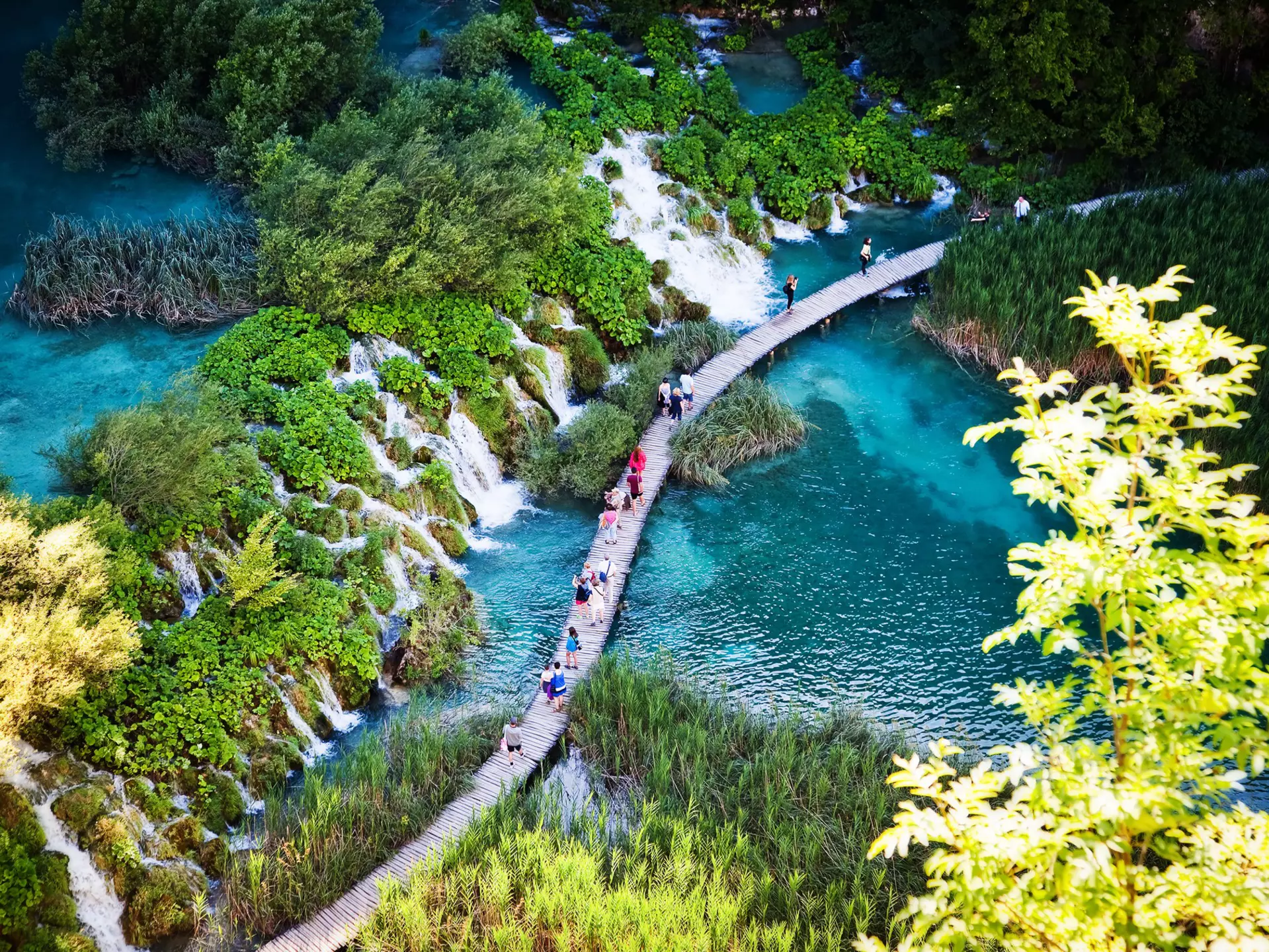 People walk on a pathway over water past a series of waterfalls.