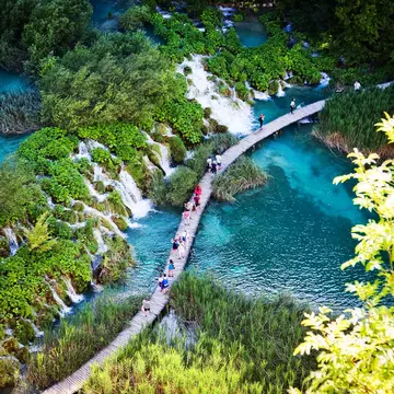 People walk on a pathway over water past a series of waterfalls.