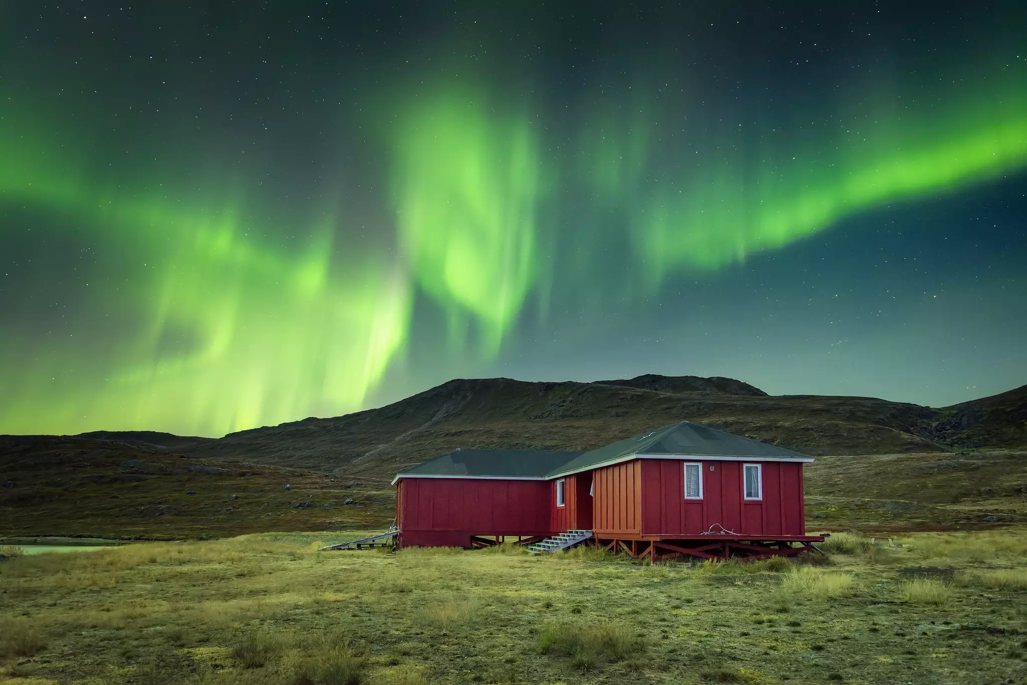 The northern lights shine over a red wooden cottage in a open field.