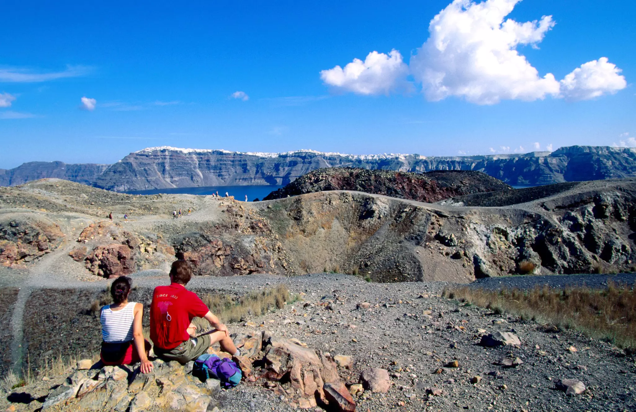 Two hikers sit on rocks in a rocky landscape looking toward peaks on the other side of blue water.