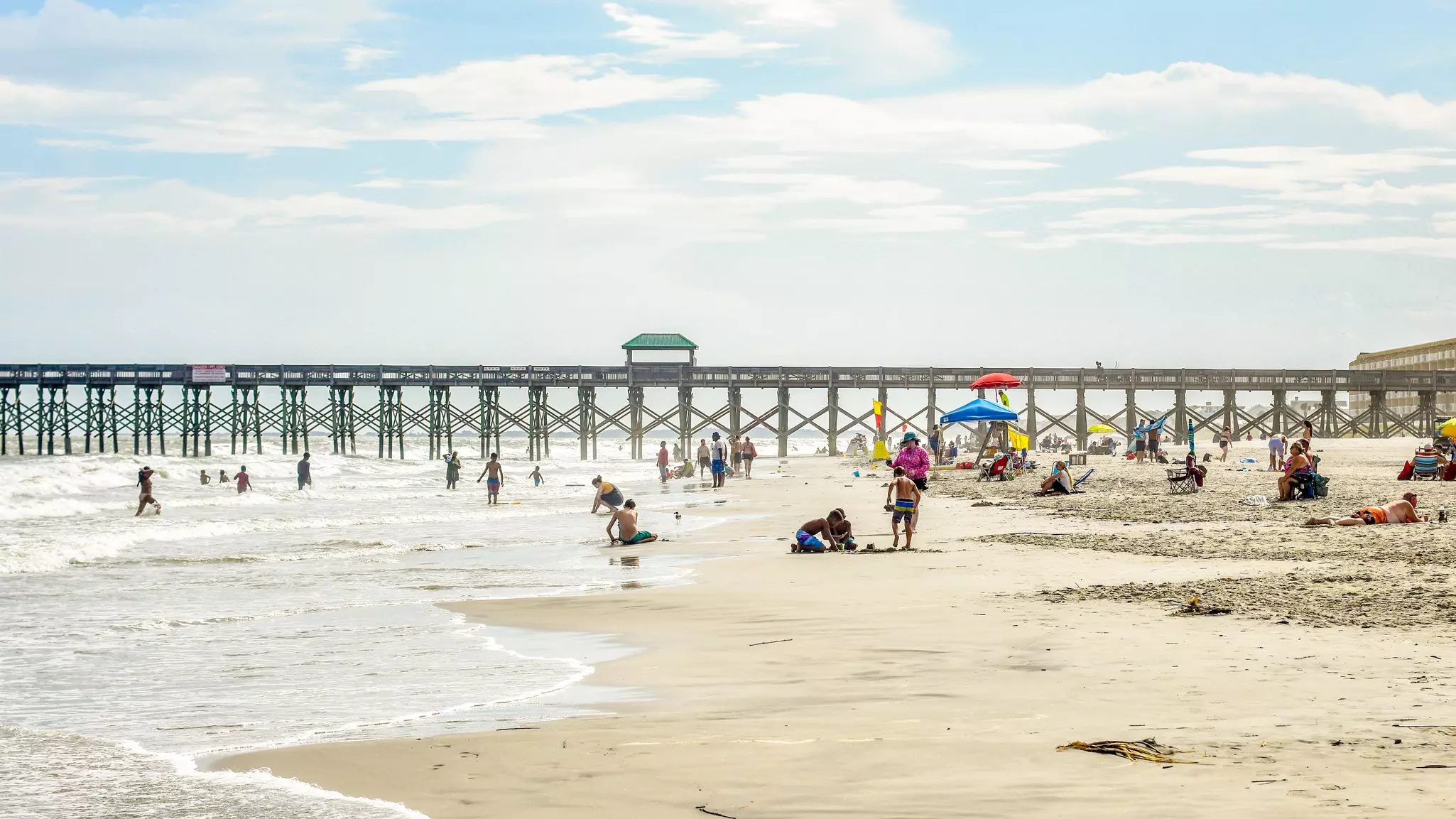 People are pictured on a beach in front of a long fishing pier.