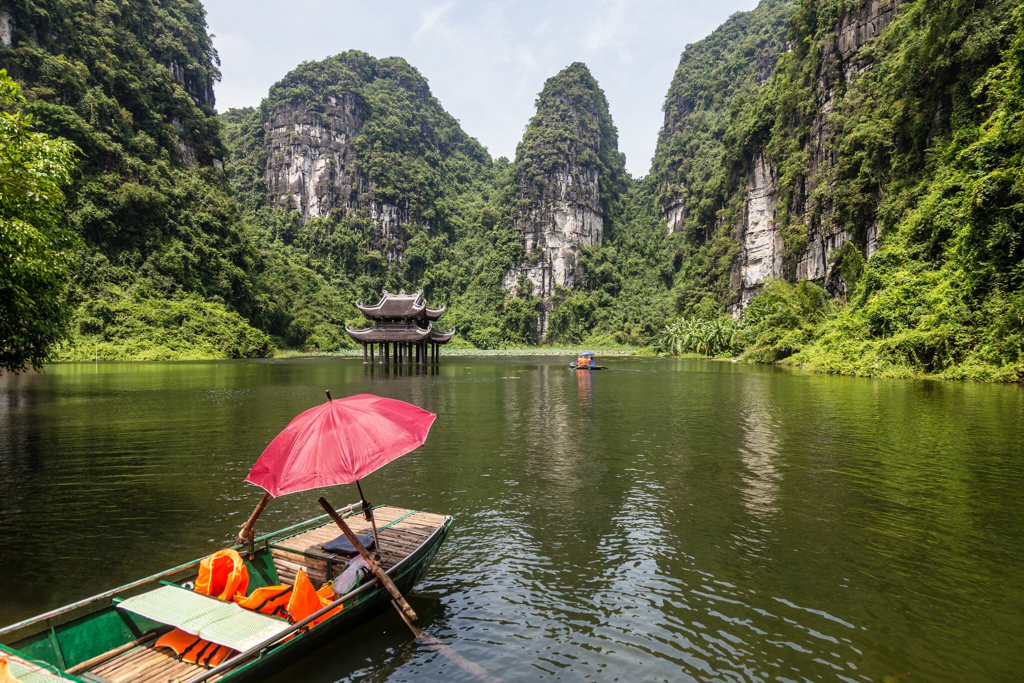 Empty sampan with a red umbrella surrounded by steep limestone cliffs and with a small temple in the background.