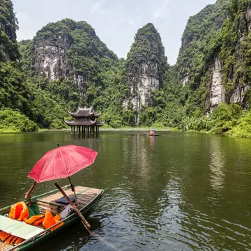 Empty sampan with a red umbrella surrounded by steep limestone cliffs and with a small temple in the background.