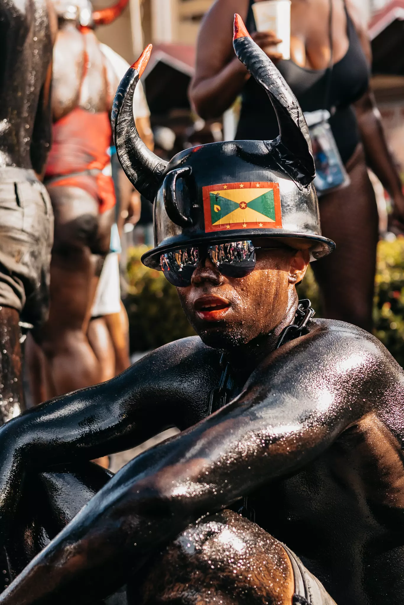 A man covered in black oil wears shades, a helmet with horns and the Grenada flag pasted to the front sits on the road in Grenada during Jab Jab for Spicemas.
