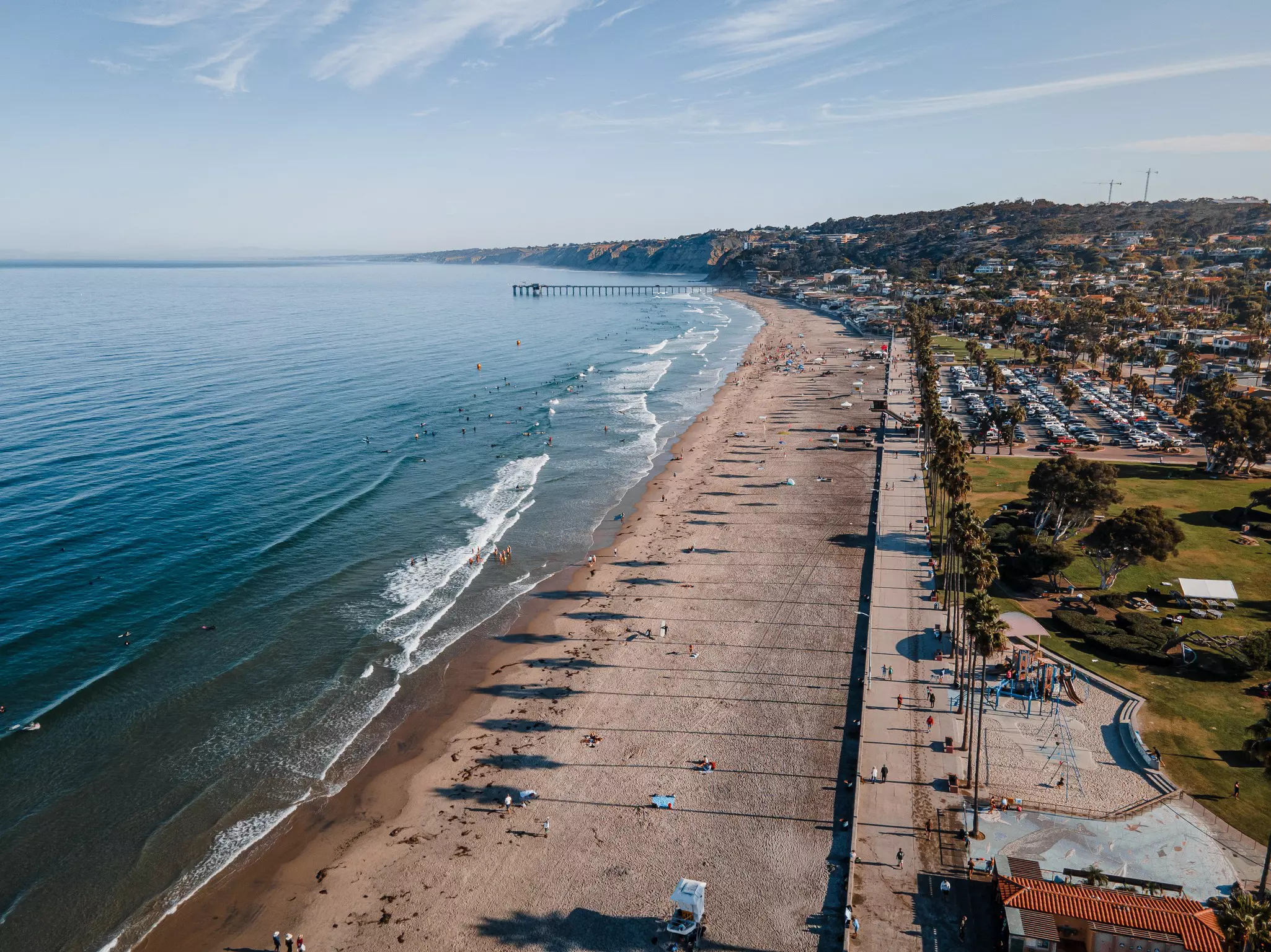 A long, wide sandy beach by blue ocean; there is a distant pier and buildings are next to the beach.