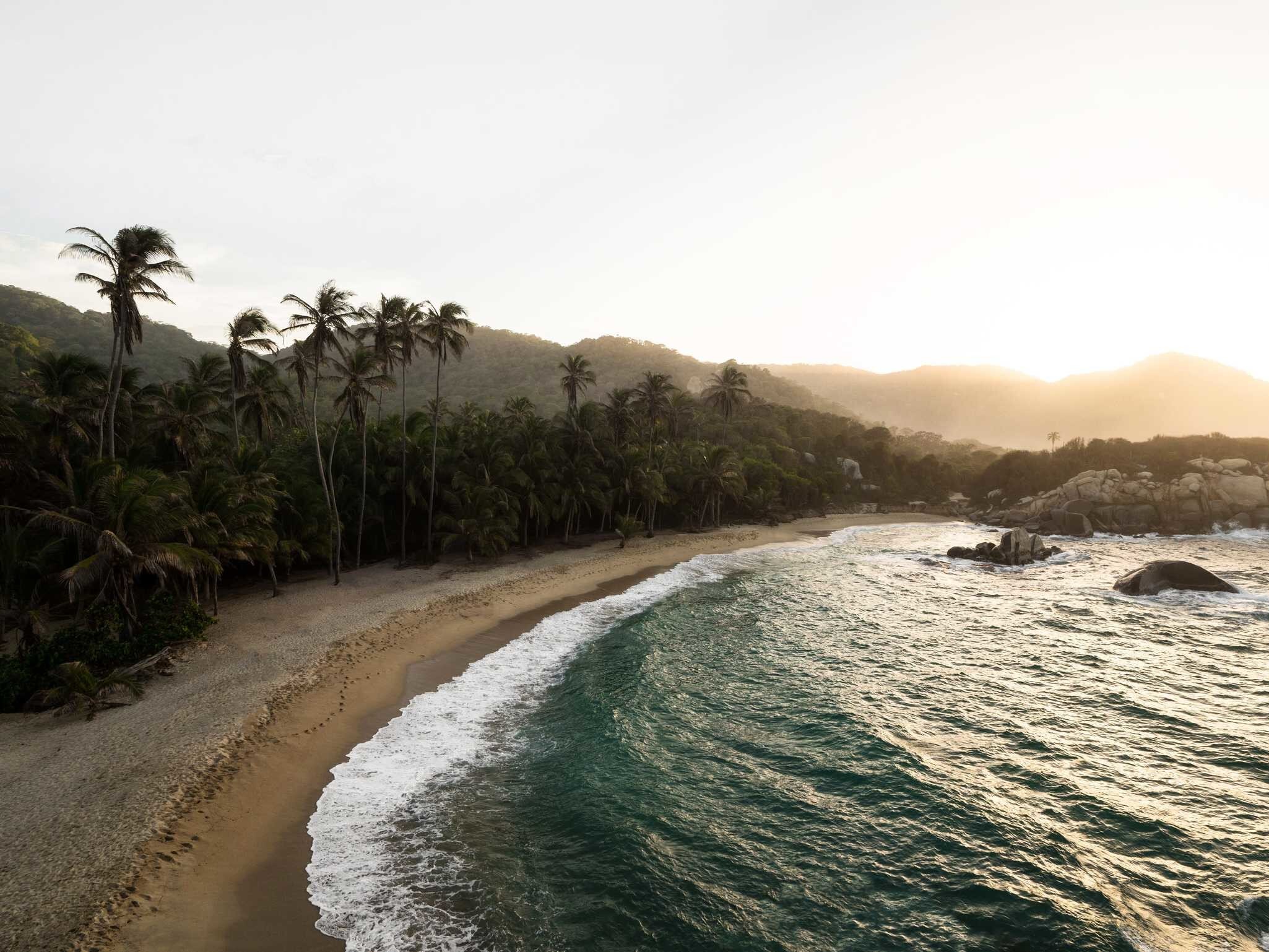 Aerial view of curved sandy beach and palm trees to the left with ocean to the right and rocks and mountains in the distance.