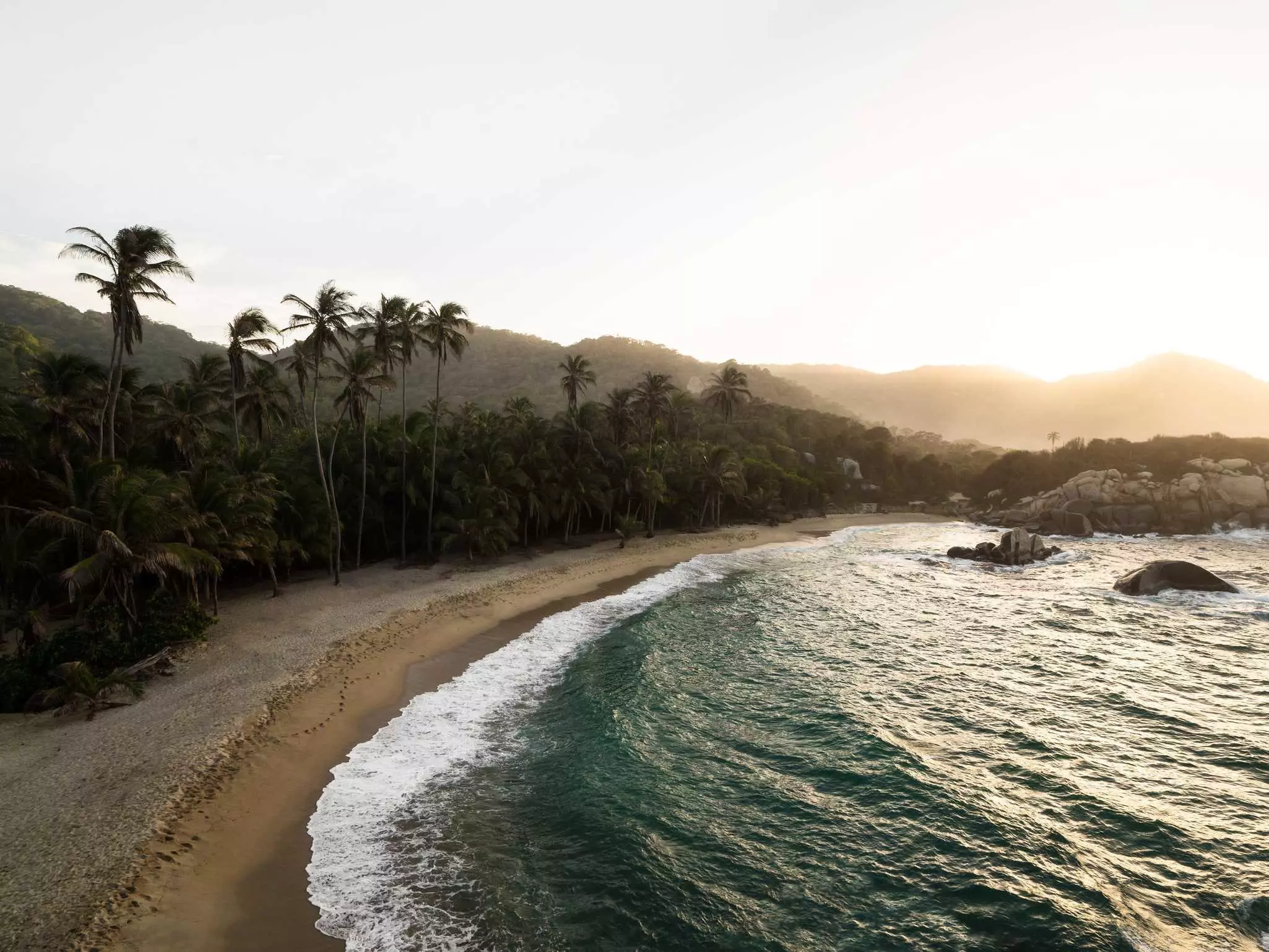 Aerial view of curved sandy beach and palm trees to the left with ocean to the right and rocks and mountains in the distance.