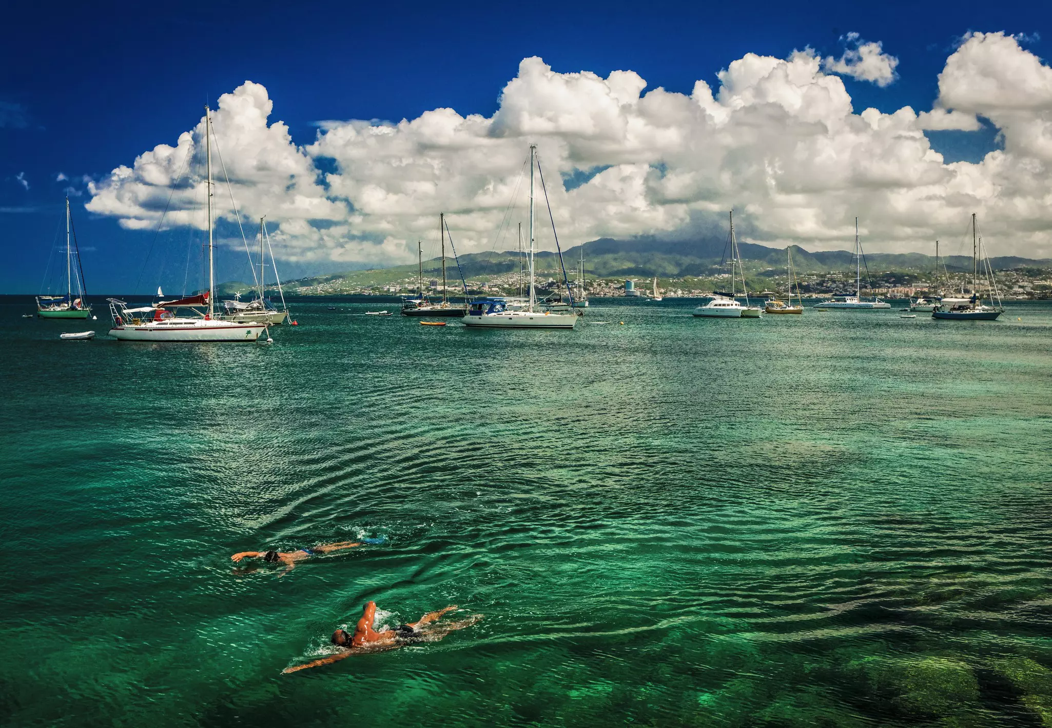 Two people swim through the water in a bay filed with sailboats. Clouds hugging green hills are visible in the distance.