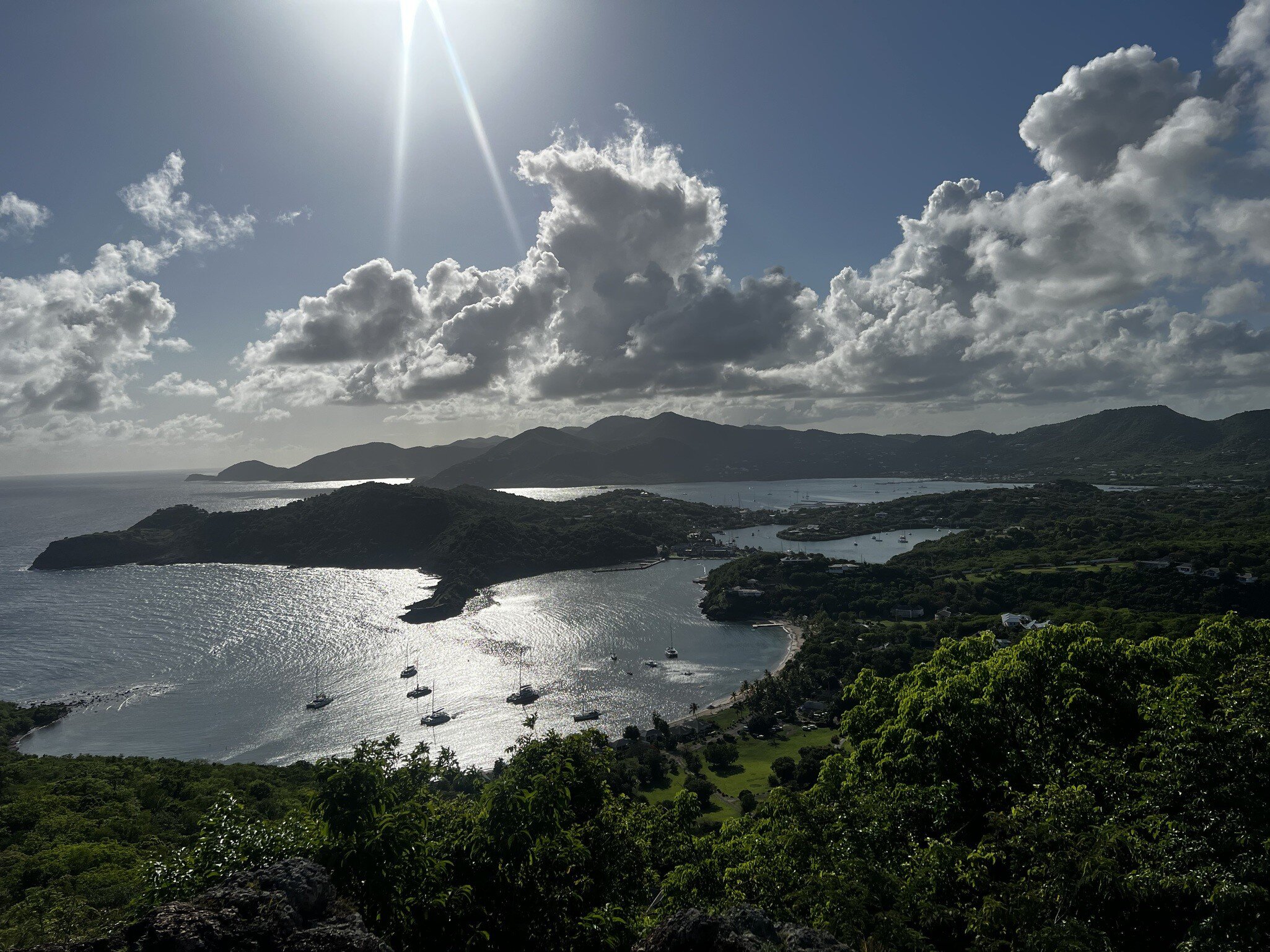 A high viewpoint over two sheltered harbours full of moored yachts, green headlands and hills stretching to the horizon under backlit cloud.