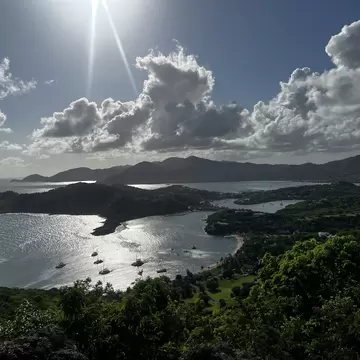 A high viewpoint over two sheltered harbours full of moored yachts, green headlands and hills stretching to the horizon under backlit cloud.