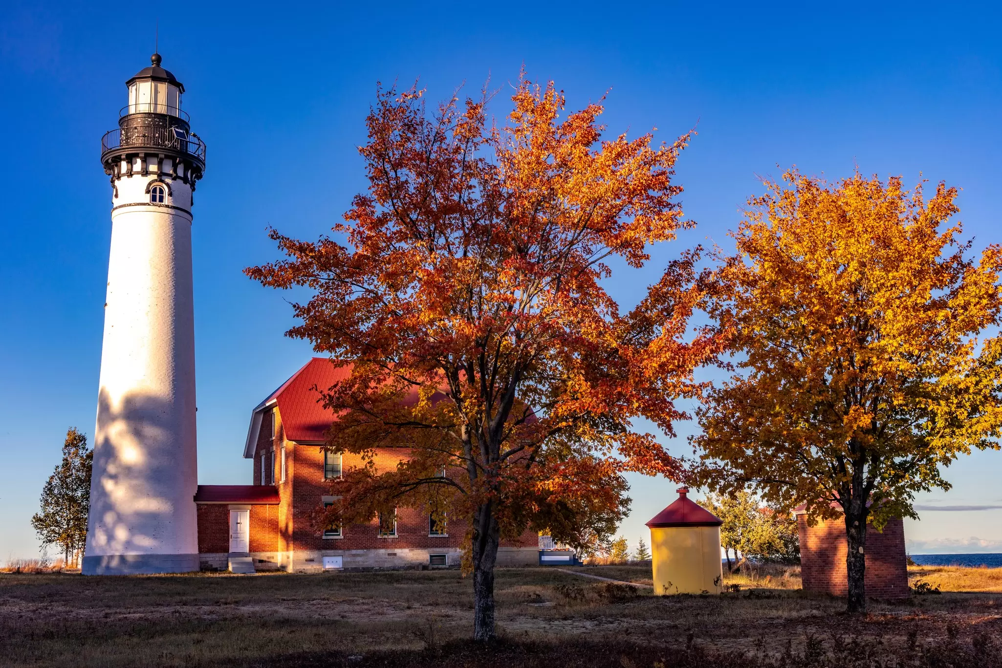 A white lighthouse is next to trees with orange leaves under blue sky.