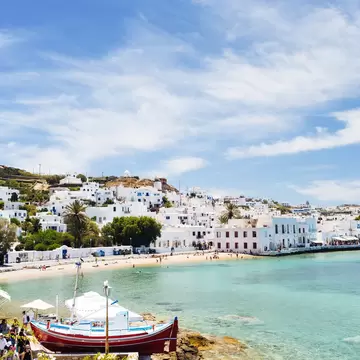 A boat sits in a turquoise harbor with whitewashed Greek buildings in the background. 