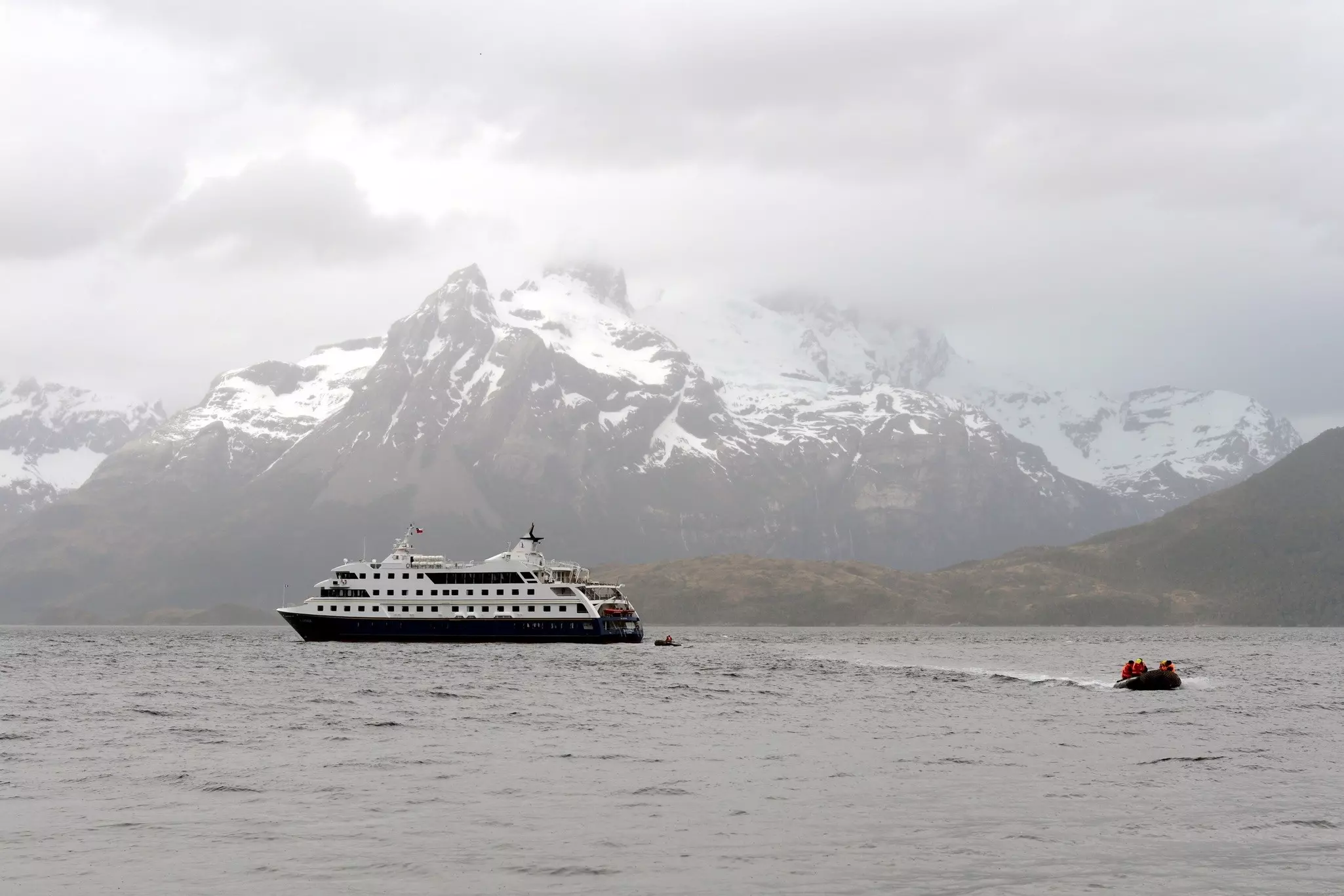 A tender near a larger boat in a waterway surrounded by misty snow-capped peaks.