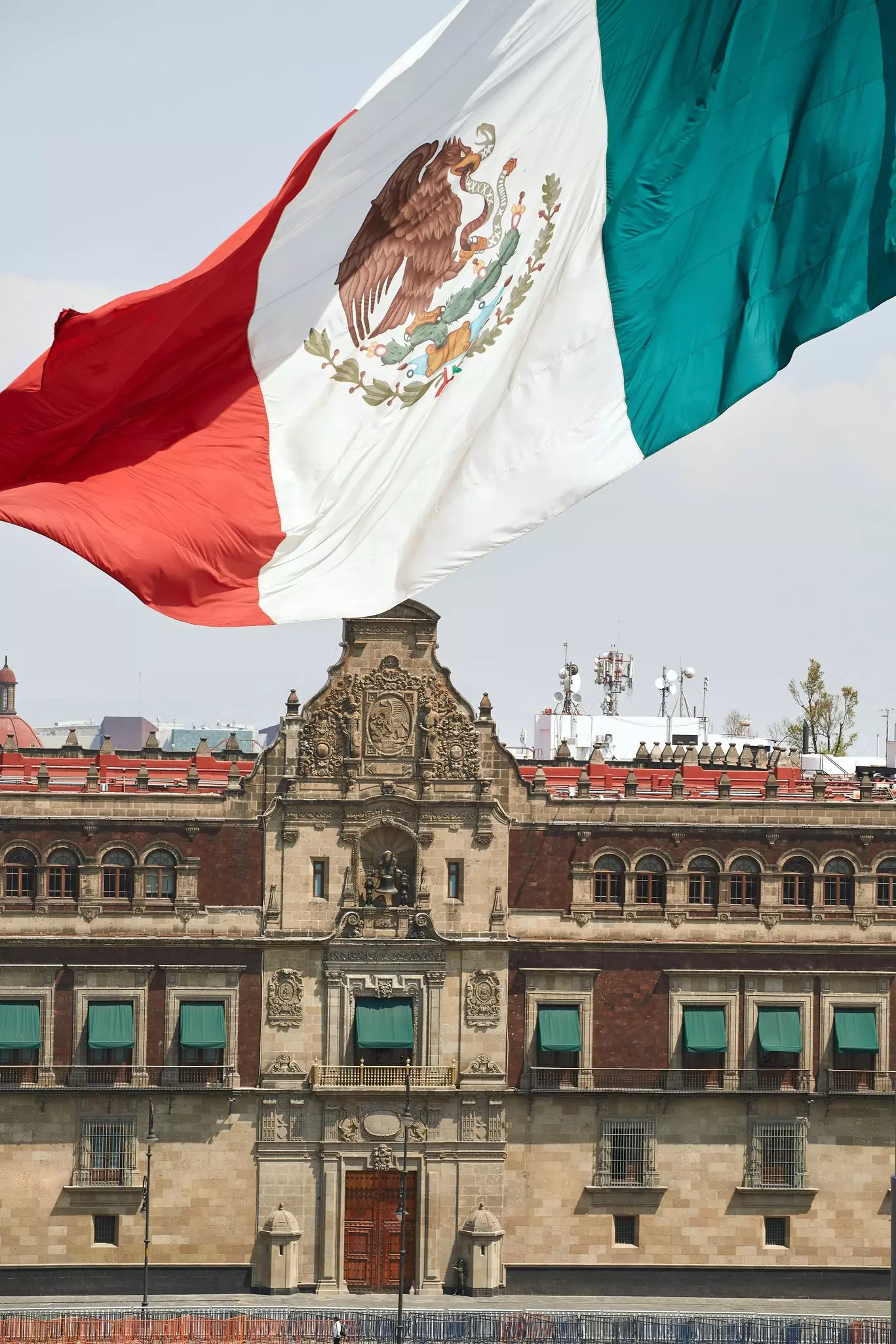 The Mexican flag – with vertical single red, white and green stripes and a crest in the middle – waving in the wind in front of the Palacio Nacional, an earth-toned building with green awnings over its second-level windows