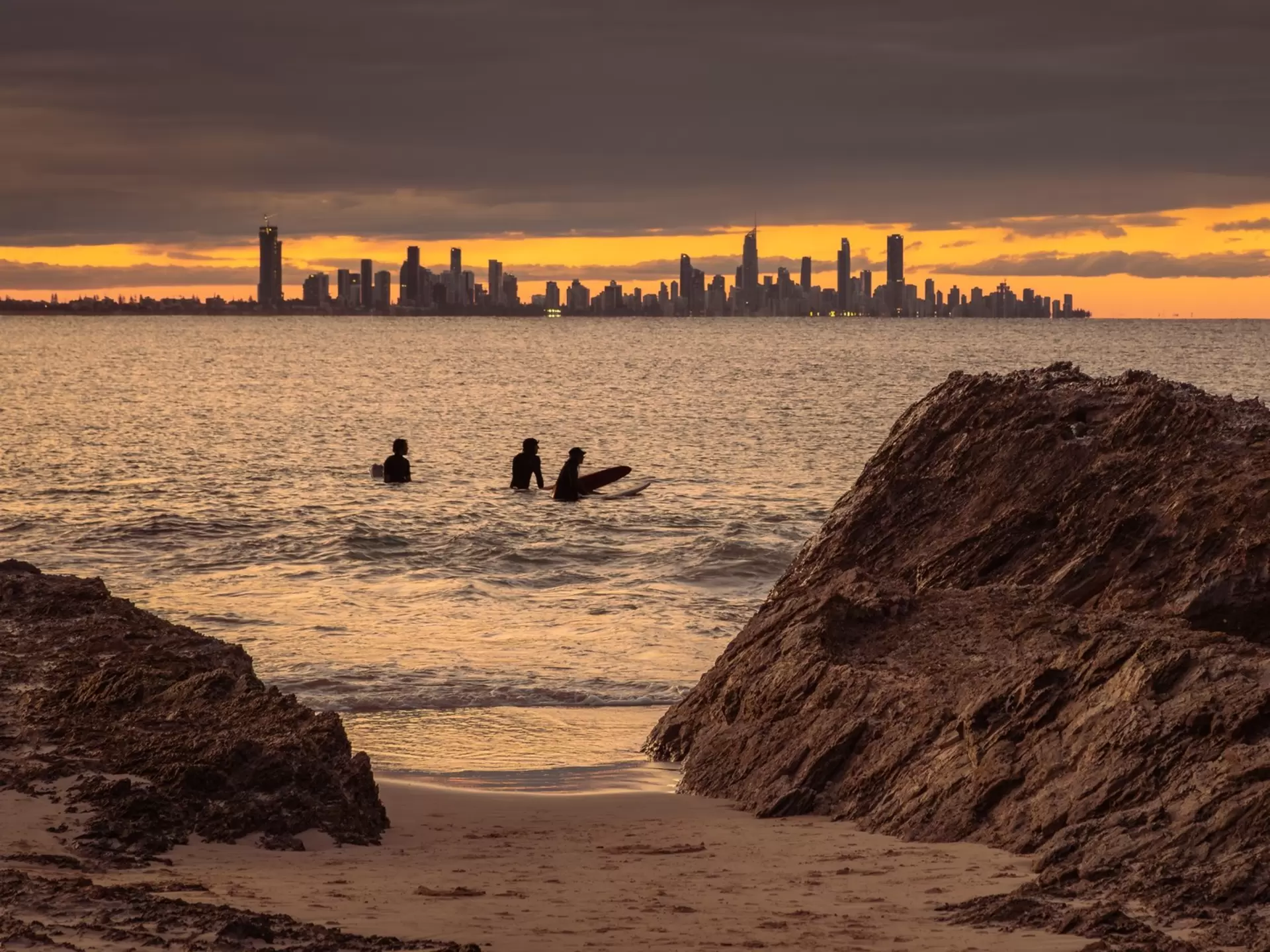 Surfers in the waters in silhouette with a city in the distance under darkening skies