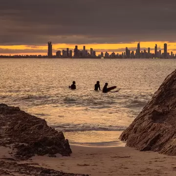 Surfers in the waters in silhouette with a city in the distance under darkening skies