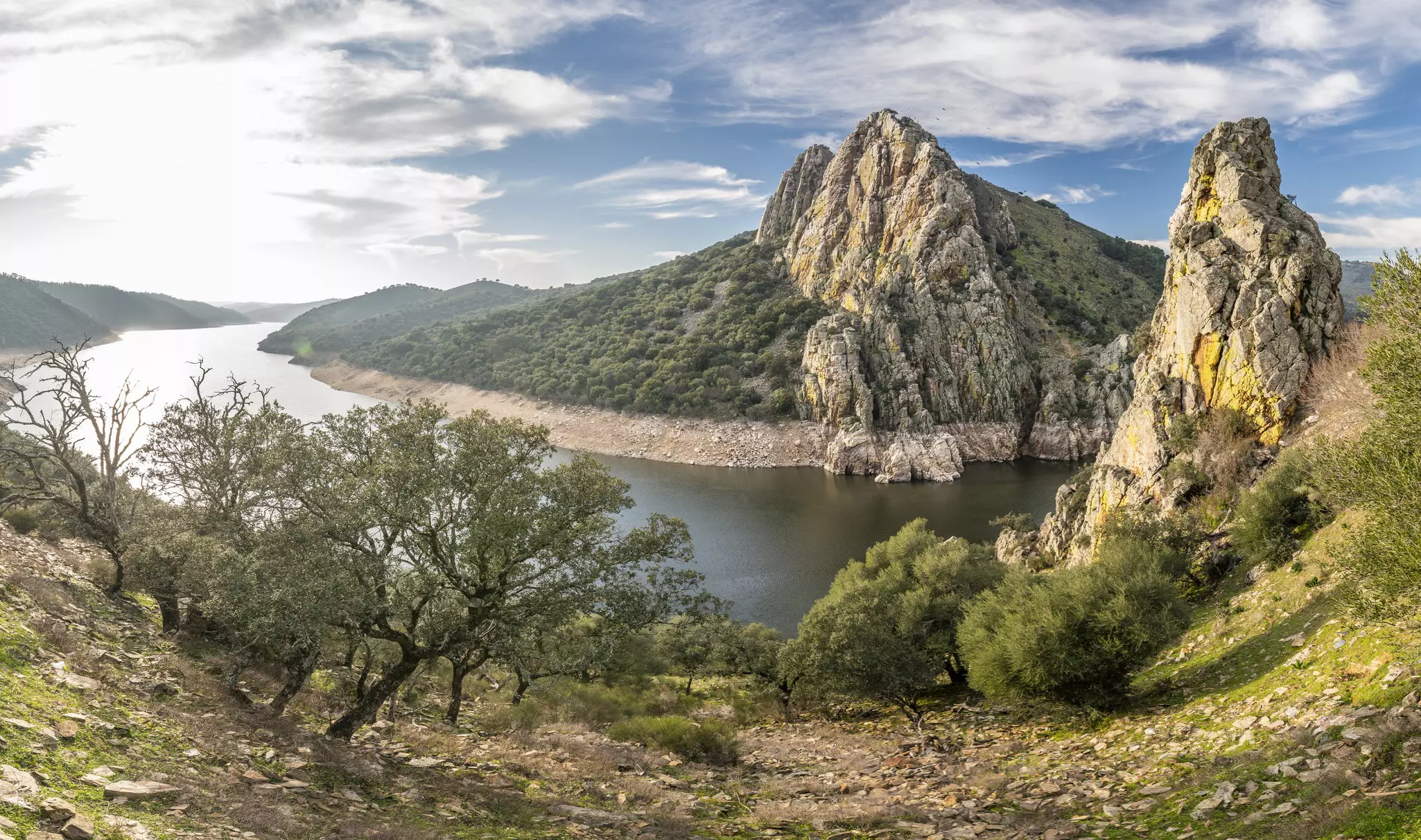 Rocky and dry landscape with olive trees and greenery overlooking a river with blue sky