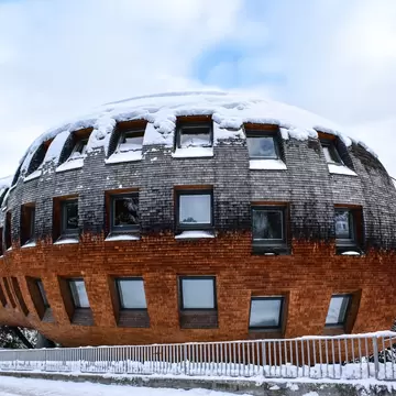Egg-shaped building clad in wooden shingles on stilts, snow on its roof and mountains behind, in St Moritz