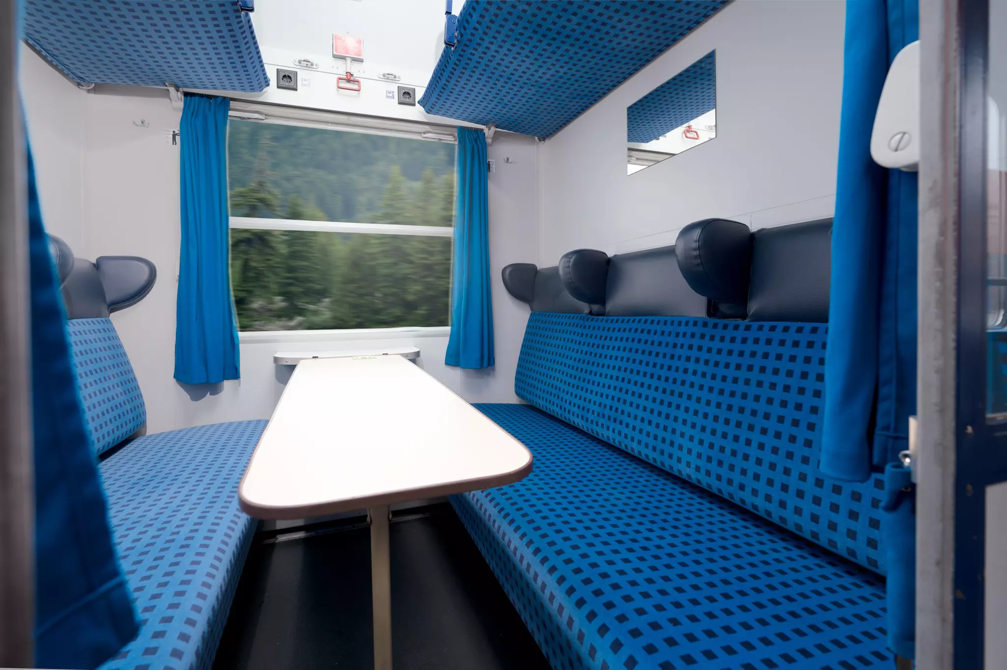 Couchette train compartment with two blue sofa seats, a white table in between them, and a window looking out to trees whizzing past.