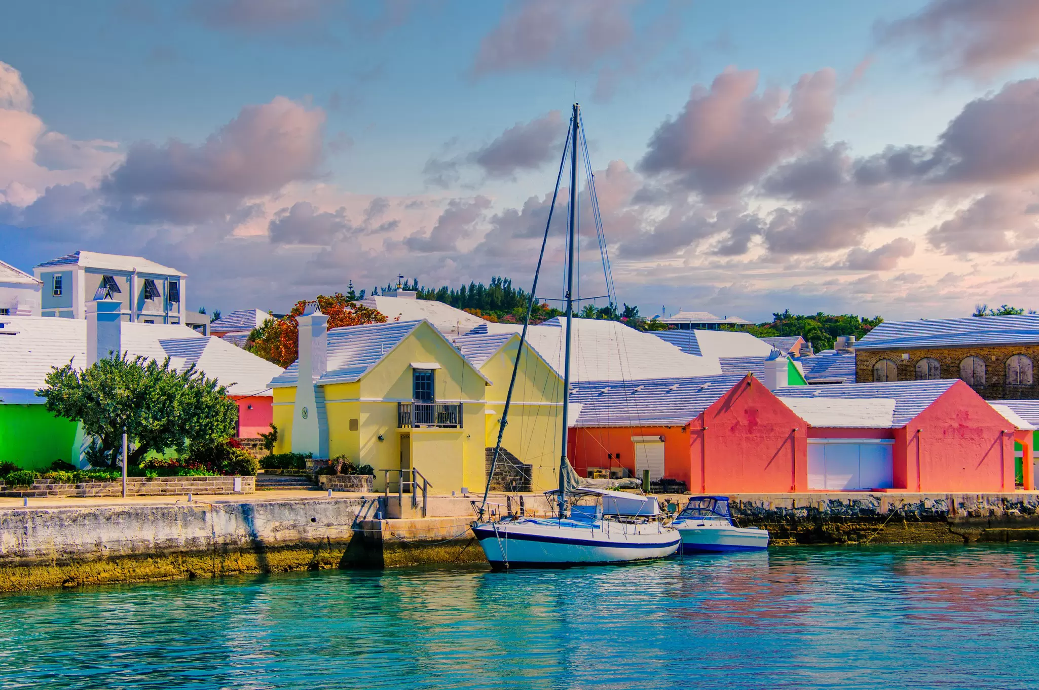 A sailboat is moored by a dock lined by pastel-colored warehouses.