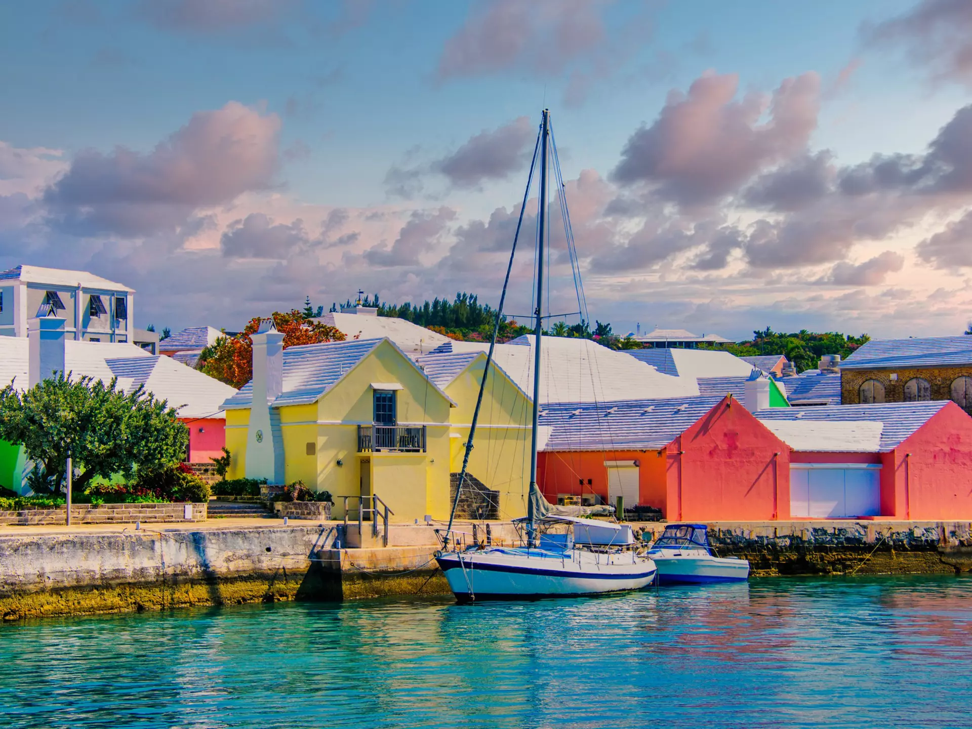 A sailboat is moored by a dock lined by pastel-colored warehouses.