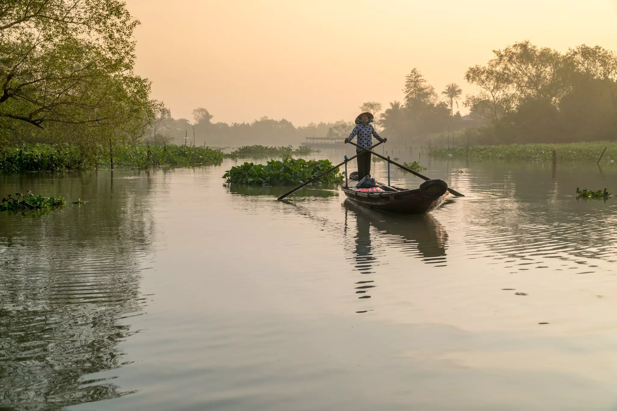 A Vietnamese woman rowing a wooden boat along a river in hazy light.