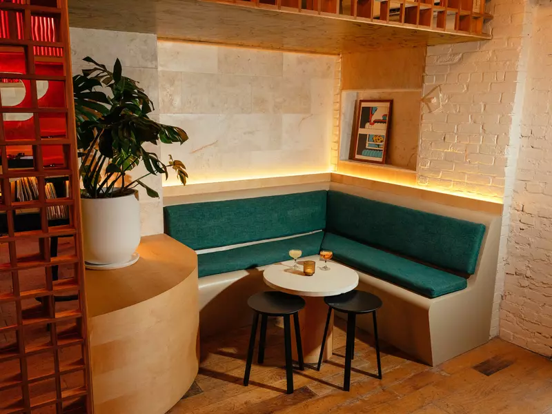 A beige corner booth with green padding faces a small round white table, under which are two black stools. There are three drinks on the table, and a plant on a shelf above the booth.