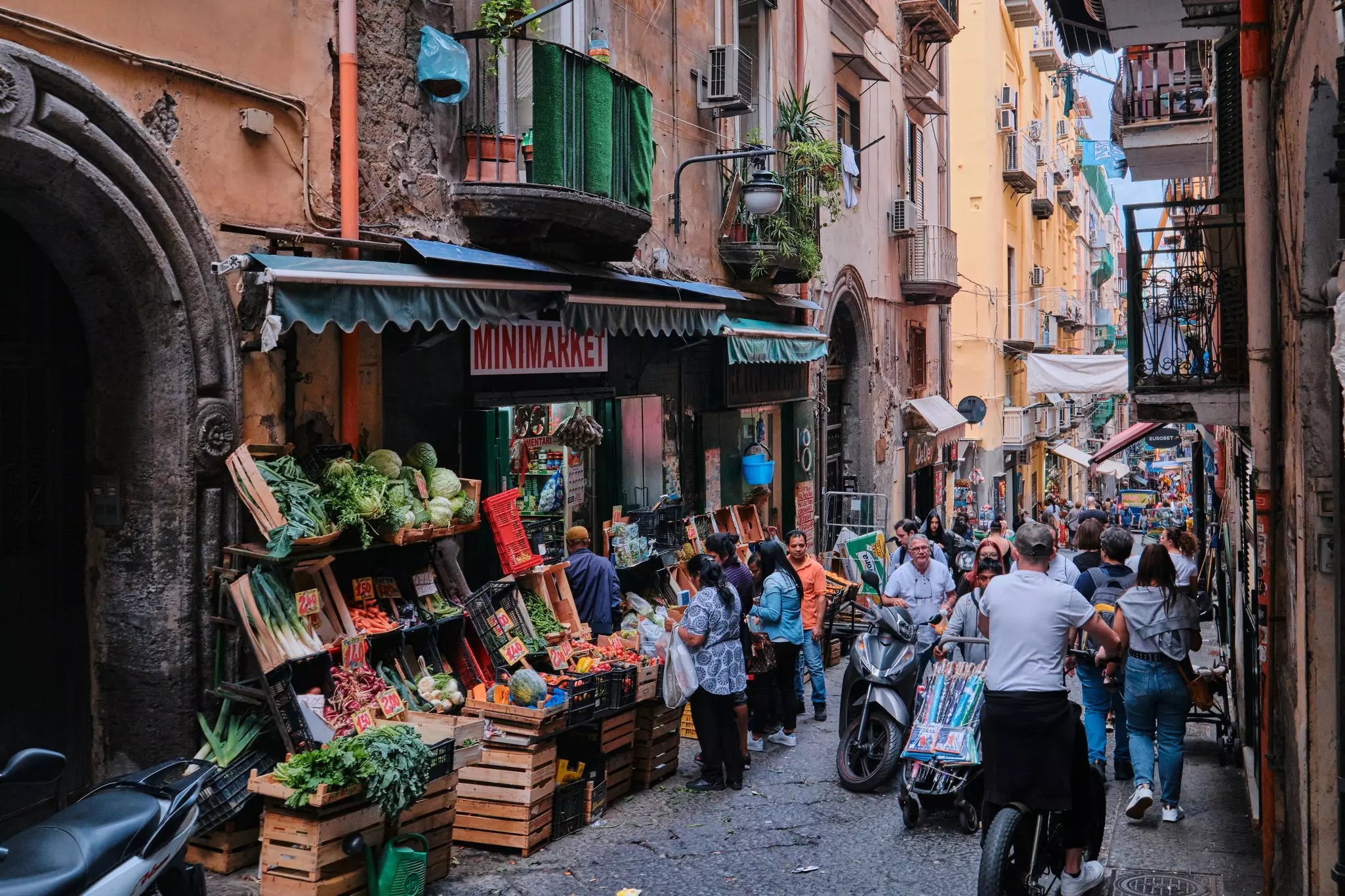 A busy narrow street with pedestrians, scooters and bikes all vying for space.