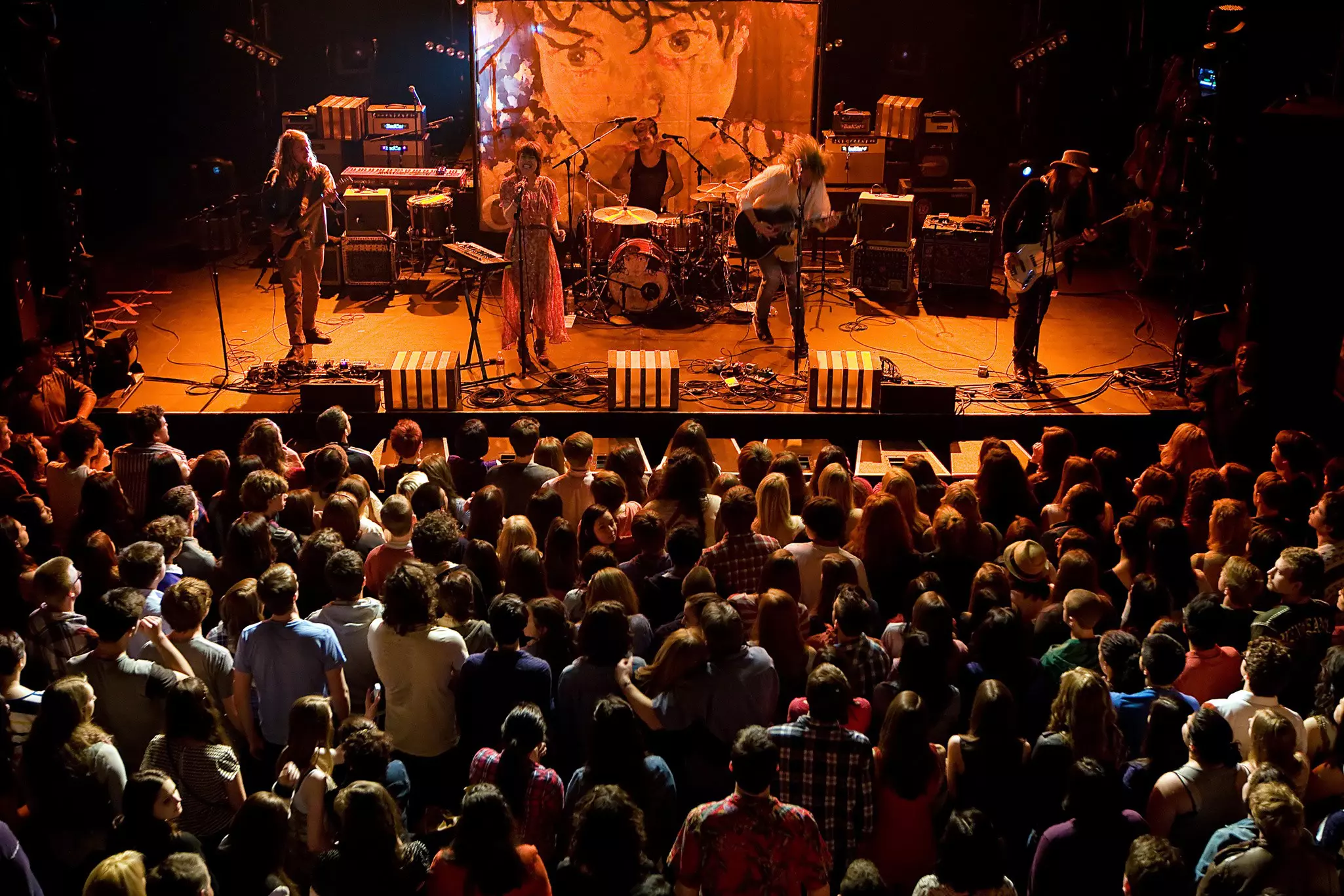 An Indie rock band perform on stage, bathed in orange light, in front of a packed standing audience.