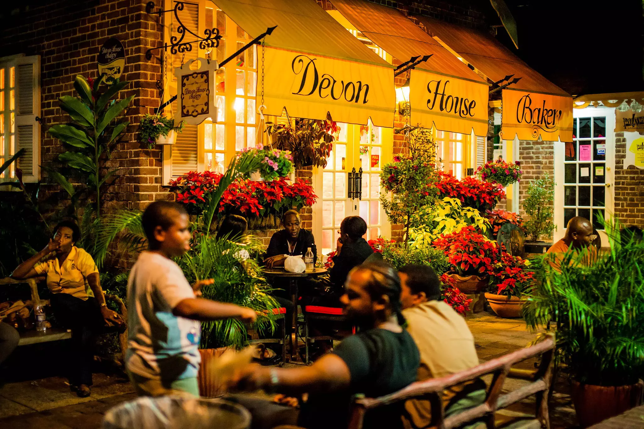 People sit at tables on a pretty flower-lined terrace outside a place with "Devon House Bakery" written on its yellow awning.