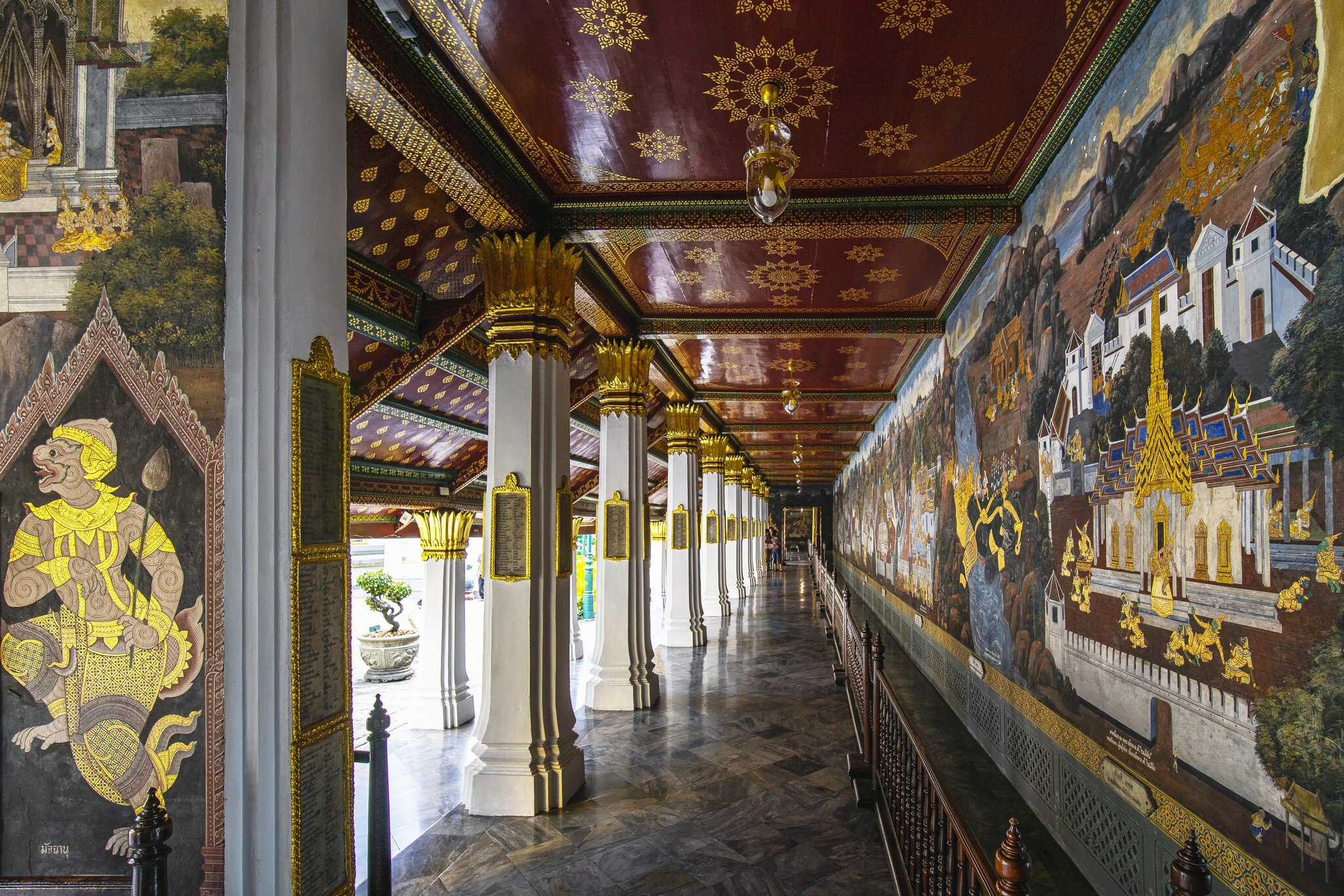 A hallway surrounding an open courtyard with elaborate, gilded pillars on one side and the brightly colored and gilded Ramakian Murals at Wat Phra Kaew temple, Bangkok