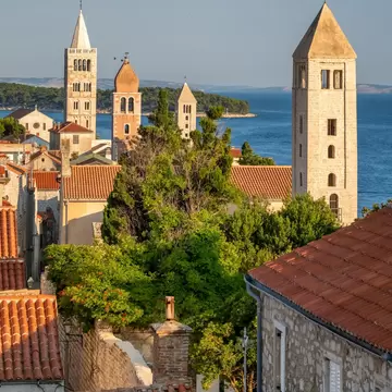 Four stone bell towers above terracotta roofs and trees in Rab town, with blue sea and a wooded island behind.