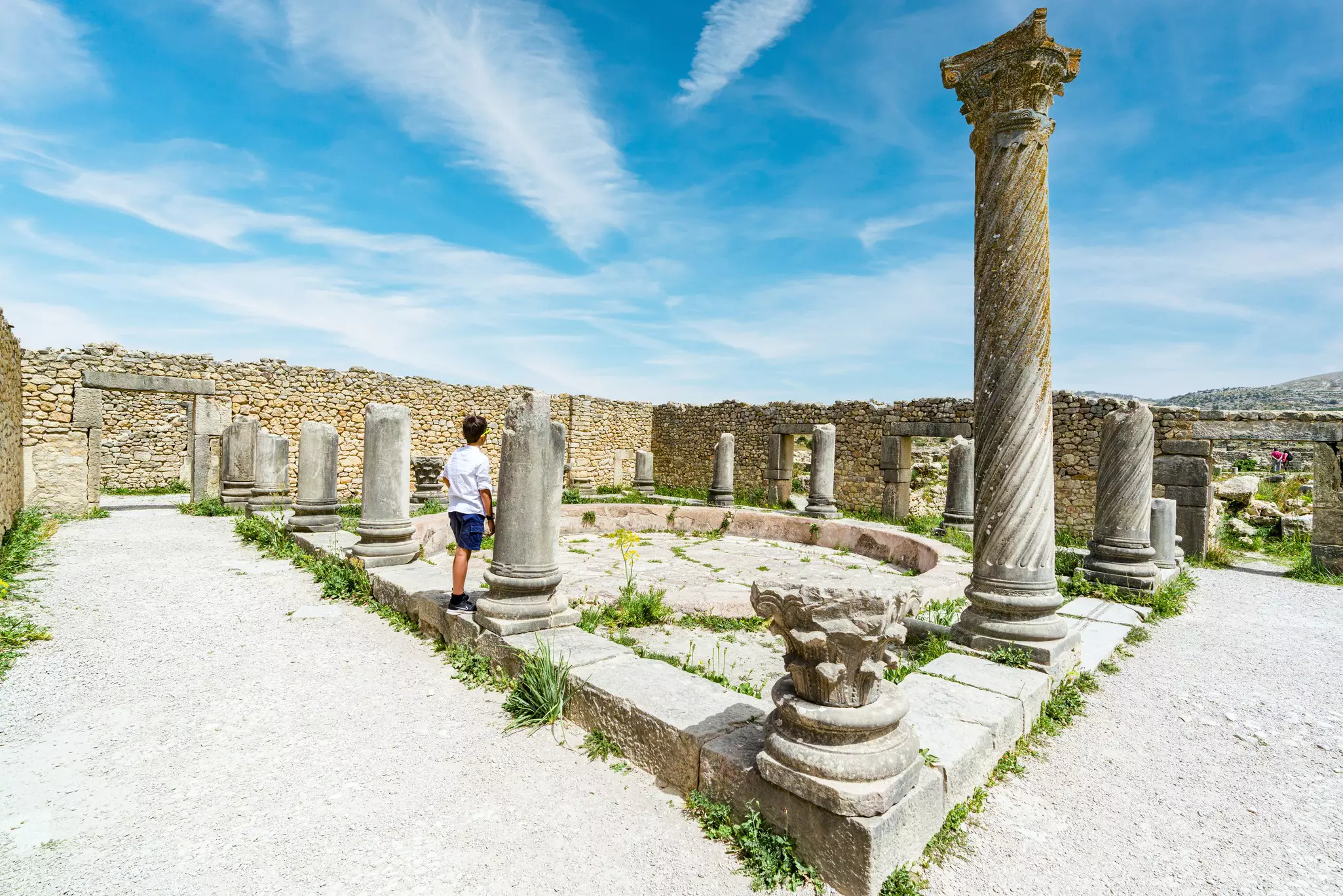 A young boy aged around 10 years old stands near an ancient Roman column looking at an archaeological site.