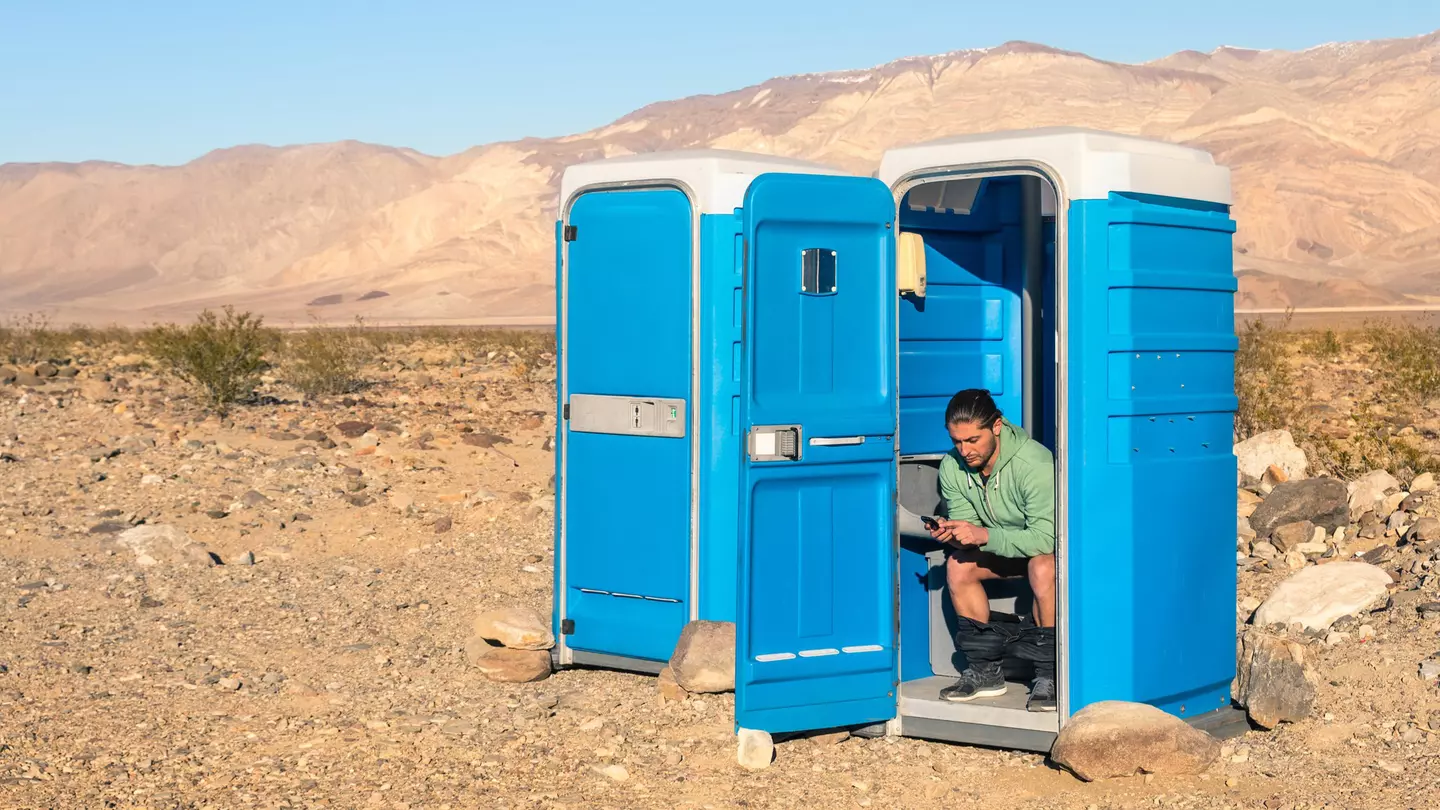 A man sits in a blue portable toilet with the door open; there is another loo next to his with the door closed. It's in Death Valley so that background is very barren.