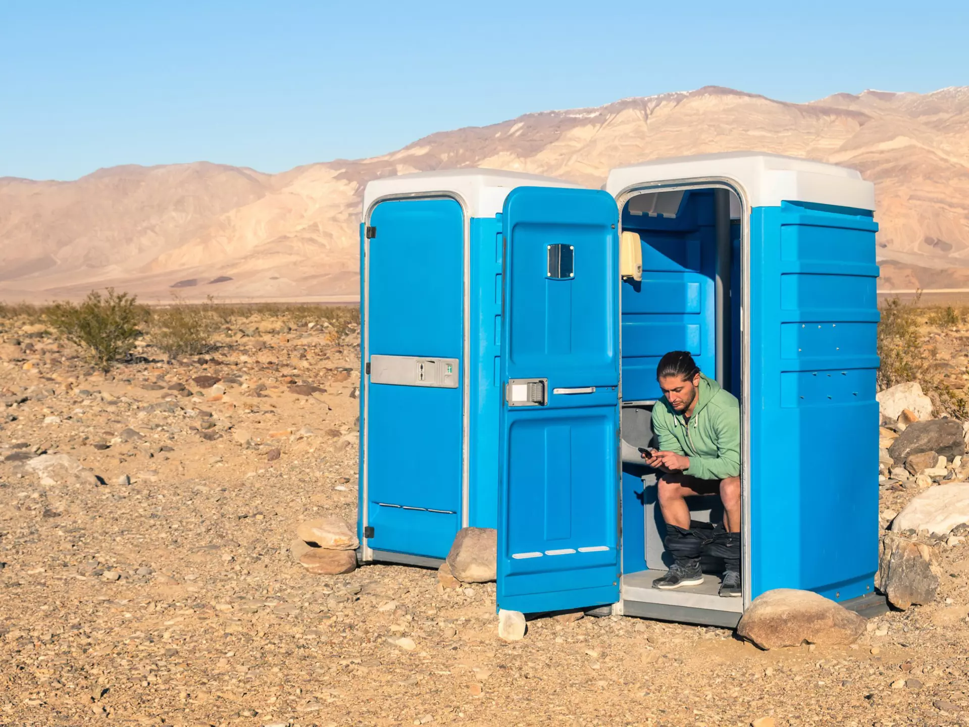 A man sits in a blue portable toilet with the door open; there is another loo next to his with the door closed. It's in Death Valley so that background is very barren.