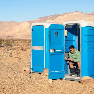 A man sits in a blue portable toilet with the door open; there is another loo next to his with the door closed. It's in Death Valley so that background is very barren.