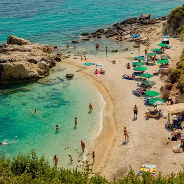 People swim in clear waters that are dotted with green parasols and backed by a forest.